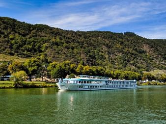 White river cruise ship MS Switzerland sailing on calm green water with forested hills and village buildings in the background under blue sky.