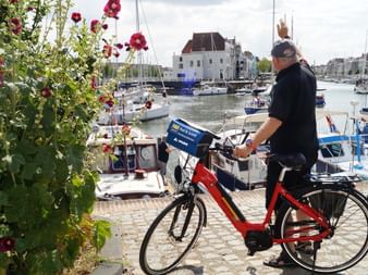 Mann mit rotem Fahrrad am Hafen von Middelburg, umgeben von Segelbooten und historischen Gebäuden. Rote Blumen blühen im Vordergrund.