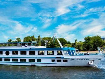 Weißes Flusskreuzfahrtschiff MS Switzerland fährt auf ruhigem Wasser bei blauem Himmel, Bäume und Grün am Ufer im Hintergrund sichtbar.
