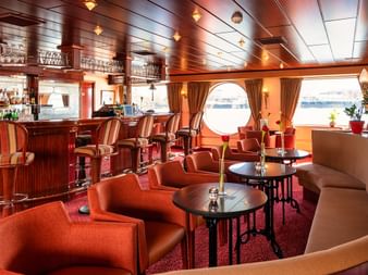 Elegante Salon-Bar auf der MS Switzerland mit orangen und beigen Sitzgelegenheiten, dunkler Holzverkleidung, runden Bullaugen und einer Theke.