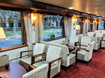 Interior of MS Classica salon with rows of white armchairs, red carpet, large windows with curtains, and warm lighting. Trees visible outside.