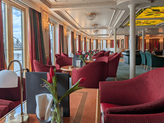 Eleganter Salon auf dem Schiff Casanova mit rot gepolsterten Stühlen, runden Tischen mit Tulpen, großen Fenstern, weißen Säulen und Zierdecke.