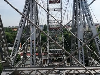Blick durch die Stahlkonstruktion eines Riesenrads mit sich kreuzenden Metallträgern und Kabeln, darunter grüne Bäume und Gebäude.