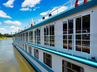 White river cruise ship MS Switzerland with turquoise trim and large windows moored on a calm river under blue sky with white clouds.