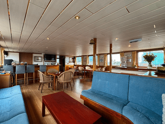 Geräumiger Salon an Bord der Clair de Soleil mit blau gepolsterten Sitzen, Holztischen, Stühlen, Barbereich und großen Fenstern mit Aussicht.