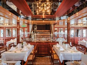 Elegant restaurant aboard MS Switzerland with white-clothed tables, wooden paddle wheel centerpiece, and brass chandelier under mirrored ceiling.