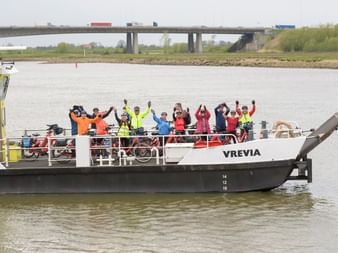 Gruppe von Radfahrern in bunten Jacken winkt vom Deck des Bootes Vrevia auf holländischem Wasserweg, Brücke im Hintergrund sichtbar.