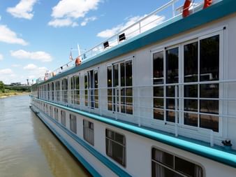 White river cruise ship with turquoise trim and large French balcony windows docked on a river under blue sky with clouds.