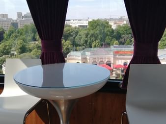 Blick durch Fenster einer Prater-Riesenradgondel mit rundem weißen Tisch und dunklen Vorhängen. Grüne Parklandschaft und historische Gebäude unten sichtbar.