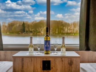 Upper deck cabin interior on MS Princess with wine bottles and glasses on wooden table, large window overlooking river and trees.