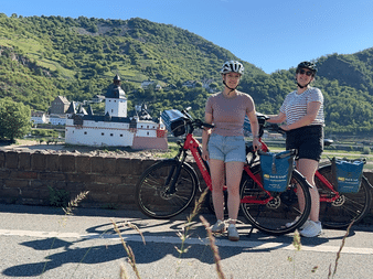 Zwei Radfahrerinnen mit roten Tourenrädern am Rheinufer mit Burg Pfalzgrafenstein und grünen Weinbergen im Hintergrund.