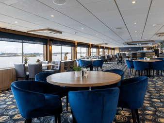 Moderne Schiffslounge mit blauen Samtstühlen um runde Tische, gemustertem Teppich und großen Fenstern mit Flussblick. Barbereich im Hintergrund.