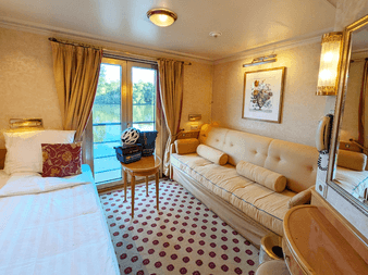 Helle Kabine mit weißem Bett, beigem Sofa, gemustertem Teppich, Balkontür mit Flussblick, goldenen Vorhängen und Spiegel auf Casanova Oberdeck.