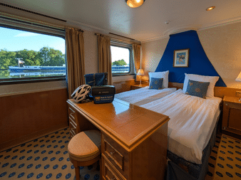 Geräumige Kabine mit Doppelbett, blauer Akzentwand, Holzschreibtisch, gemustertem Teppich und großen Fenstern mit Flussblick auf der Casanova.