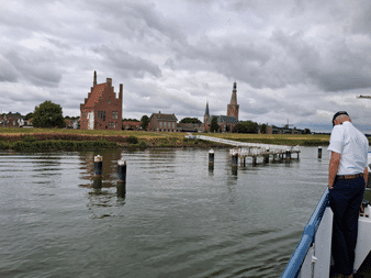 Blick vom Boot auf Medemblik Hafen mit historischem roten Backsteingebäude, Kirchtürmen und Holzsteg. Person in weißem Hemd steht auf Bootsdeck.