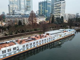 MS Classica river cruise ship with white hull and orange stripe moored at urban waterfront. Modern skyscrapers and bare trees visible in background.