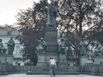 Person betrachtet das Martin-Luther-Denkmal in Worms mit zentraler Luther-Statue, umgeben von sitzenden Figuren auf Sockeln.