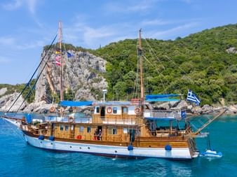 Traditionelle hölzerne Segelyacht Panagiota mit weißem Rumpf und Holzaufbau vor Anker in türkisfarbenem Wasser vor griechischer Küste.