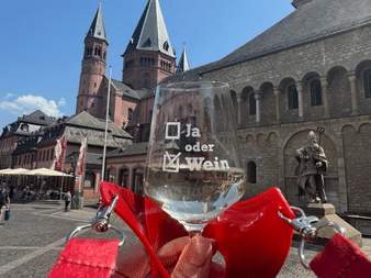 Hand hält Weinglas mit deutschem Text 'Ja oder Wein' und rotem Bändchenhalter am Mainzer Domplatz mit historischen Domtürmen im Hintergrund.