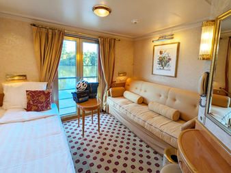 Helle Kabine mit weißem Bett, beigem Sofa, gemustertem Teppich, Balkontür mit Flussblick, goldenen Vorhängen und Spiegel auf Casanova Oberdeck.