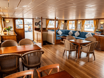 Heller Salon an Bord der Clair de Soleil mit Holzboden, Korbstühlen, blauem Sofa, goldenen Vorhängen und großen Fenstern mit Blick auf das Deck.