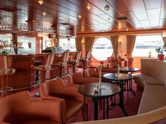 Elegante Salon Bar auf dem Schiff Switzerland mit Holztheke, Ledersesseln, runden Tischen und runden Bullaugen mit Blick aufs Wasser.