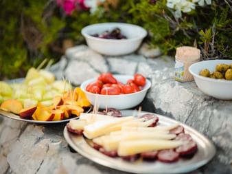 Griechische Delikatessen auf Steinoberfläche: Käse-Fleisch-Platte, Kirschtomaten, Oliven, frische Früchte und Wein in natürlicher Gartenumgebung.