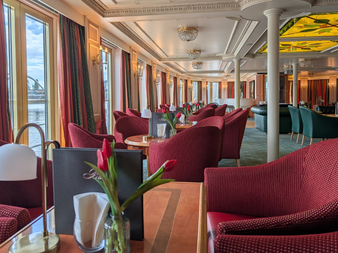 Elegant salon on the Casanova ship with red upholstered chairs, round tables with tulips, large windows, white columns, and decorative ceiling.
