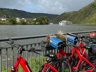 Red rental bicycles parked along a railing overlooking the Moselle River in Beilstein, with cruise ships, village houses, and green hills.