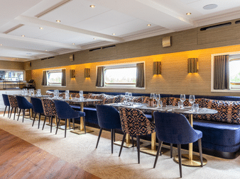 Modernes Restaurant-Interieur auf Magnifique X mit langer Tafel, blauen Samtstühlen, gemusterter Sitzbank und warmer Wandbeleuchtung unter Kassettendecke.