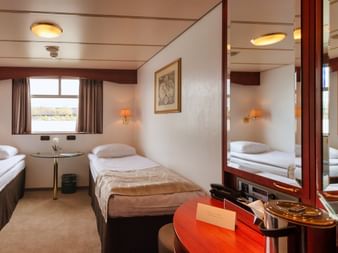 Zweibettkabine auf dem Hauptdeck der MS Switzerland mit zwei Betten, Fensterblick, Schminktisch, Spiegel, Wandlampen und beige-brauner Einrichtung.