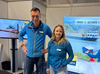 Zwei Mitarbeiter in blauen Jacken an einem Messestand mit Bildschirmen, die Flusskreuzfahrtschiffe und Radtouren zeigen.