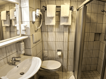 Compact bathroom on MS Classica with beige tiles, white sink, toilet, glass shower enclosure, wall-mounted hairdryer, and white towels.