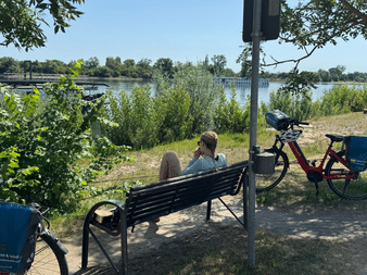 Zwei Personen sitzen auf einer Bank am Rheinufer in Nierstein. Fahrräder mit blauen Taschen stehen daneben. Grüne Vegetation und Wasser sichtbar.