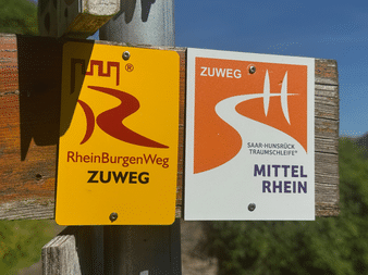 Zwei Wanderwegschilder an einem Holzpfosten: gelbes RheinBurgenWeg Zuweg-Schild und oranges Saar-Hunsrück Traumschleife Mittelrhein-Schild.