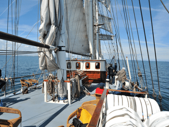 Deck der Mare Frisium mit weißen Segeln, Takelage, Korbstühlen und Passagieren. Blaues Meer und Himmel im Hintergrund.