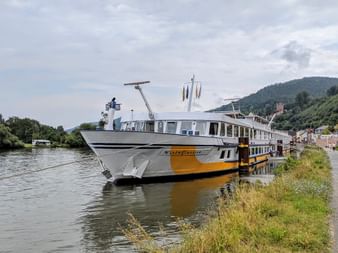 MS Classica river cruise ship with white and yellow hull moored along a green riverbank. Hills and buildings visible in background under cloudy sky.