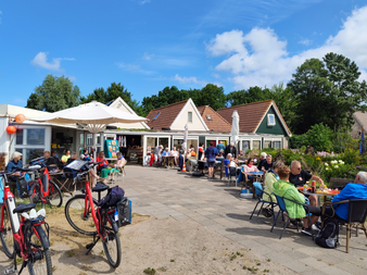 Café-Terrasse mit Radfahrern und Gästen an Tischen. Rote Fahrräder im Vordergrund, weiße Gebäude mit roten Dächern dahinter.