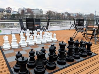 Sun deck of MS Classica with oversized chess set on wooden deck. Black folding chairs arranged around the board with river and cityscape visible.