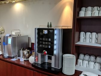 Coffee station with automatic espresso machine and water dispenser on wooden counter. White cups and plates arranged on shelves above.