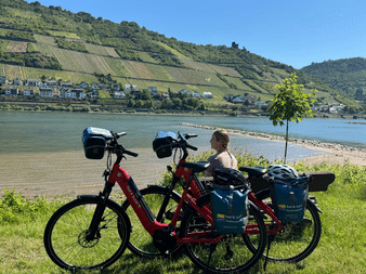 Zwei rote Tourenräder stehen im Gras am Rheinufer. Ein Radfahrer sitzt daneben. Terrassierte Weinberge bedecken den Hang am anderen Ufer.