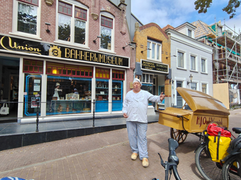 Person in weißer Bäckeruniform neben gelbem Brotwagen vor dem Bakkerijmuseum in Medemblik, mit historischen Ladenfronten im Hintergrund.