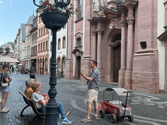 Familie auf einem Kopfsteinpflasterplatz in der Mainzer Altstadt. Zwei Kinder sitzen auf einer Bank, während ein Erwachsener mit einem roten Bollerwagen steht.