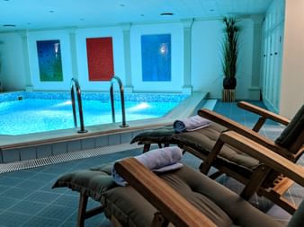 Indoor pool with blue mosaic tiles and wooden lounge chairs with towels. Three colorful water features on the wall and decorative plants.