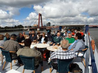 Gruppe von Passagieren an Tischen auf dem Sonnendeck des Schiffes De Nassau. Rote Brücke und Fluss im Hintergrund unter bewölktem Himmel.