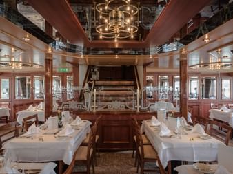 Elegant ship restaurant with white-clothed tables, wooden chairs, and a historic paddle wheel centerpiece. Mirrored ceiling and warm lighting.
