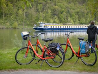 Rote Tourenräder am Donauufer geparkt mit einem blau-weißen Passagierschiff im Hintergrund, umgeben von grünem Wald.