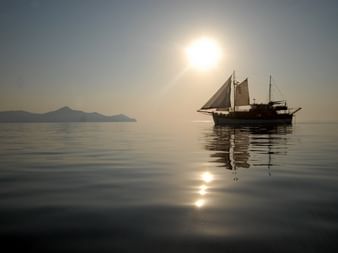 Traditionelles Segelschiff Panagiota als Silhouette vor goldenem Sonnenuntergang auf ruhigem Wasser, mit Bergen im Hintergrund.