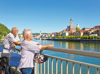 Älteres Paar mit Fahrrädern steht am Brückengeländer und blickt auf Passaus historische Altstadt mit Kirchtürmen und bunten Gebäuden an der Donau.
