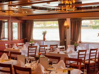 Elegantes Restaurant an Bord der MS Switzerland mit weiß gedeckten Tischen, Holzstühlen und großen Fenstern mit Blick aufs Wasser.